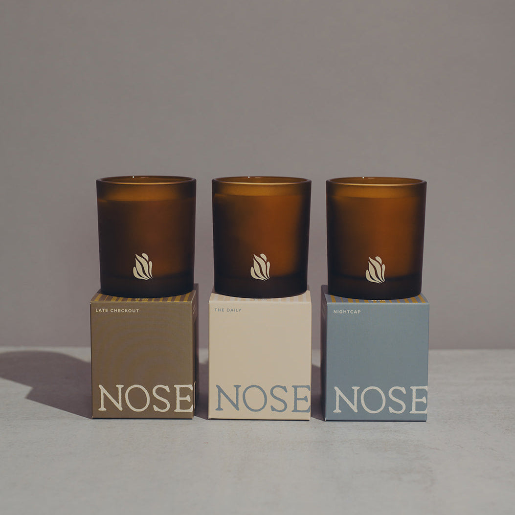 Three amber-colored candles with 'NOSE' branding on their lids, displayed on matching boxes against a neutral background.